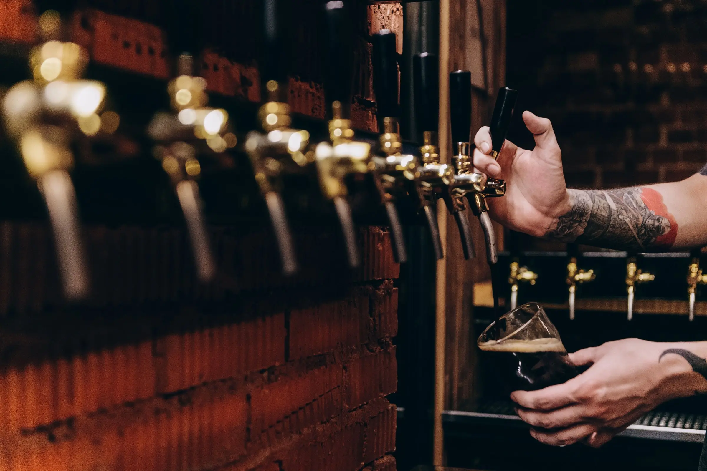 Row of brass beer taps on an exposed-brick wall, hands pouring a dark beer